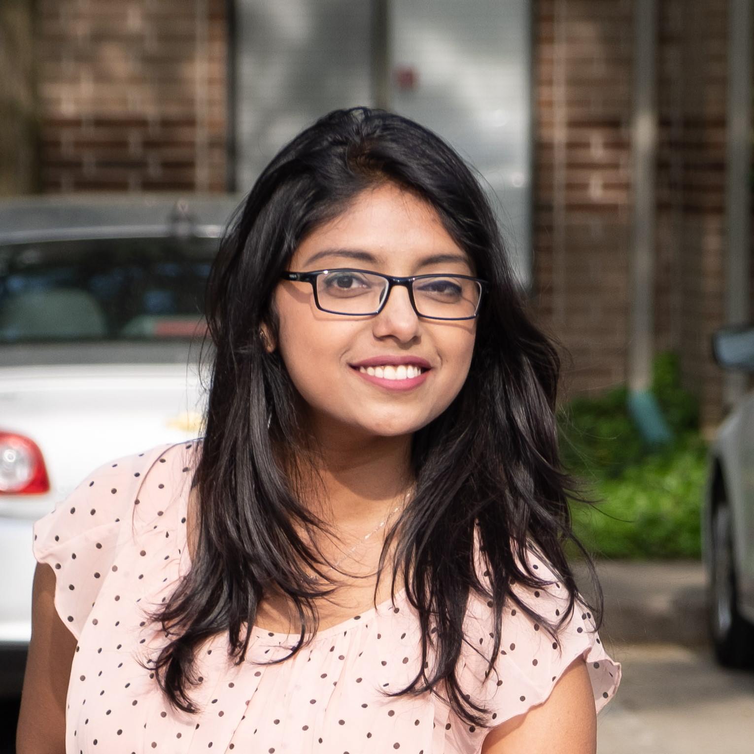 Maitraye, a young, Brown woman with black shoulder-length straight hair, is smiling at the camera. She is wearing square glasses and a peach-colored polka-dotted dress. Background shows a car in front of a red wall.