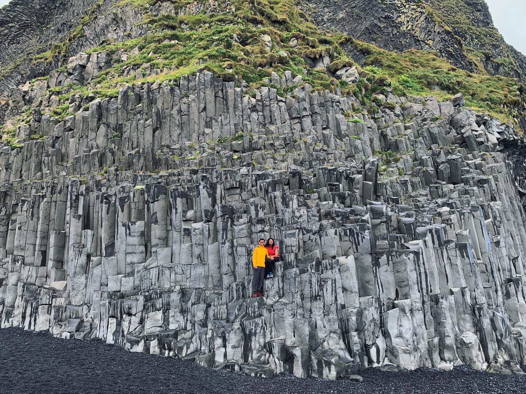Maitraye and her husband sitting on top of a basalt column in Reynisfjara black sand beach in Iceland